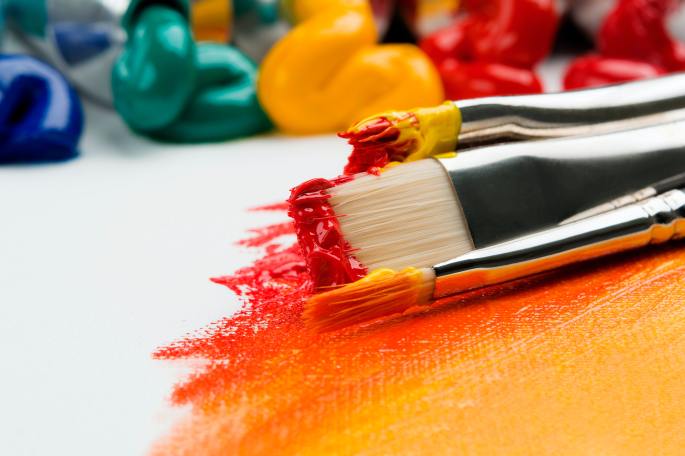 A close-up of three paintbrushes with thick acrylic paint in red and yellow shades. They are laying on a canvas with a red, orange, and yellow ombre. In the background, there are several tubes of paint with paint squeezed out, showing piles of acrylic paint in blue green, yellow, orange, and red hues.