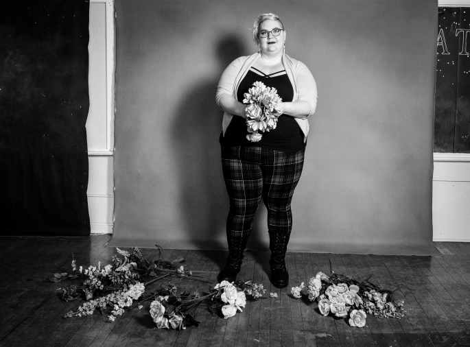 Black and white photo of Caitlin facing the camera holding a bouquet of fake flowers, with other flowers at their feet. Caitlin's face is serious and thoughtful for this blog about traumaversary.