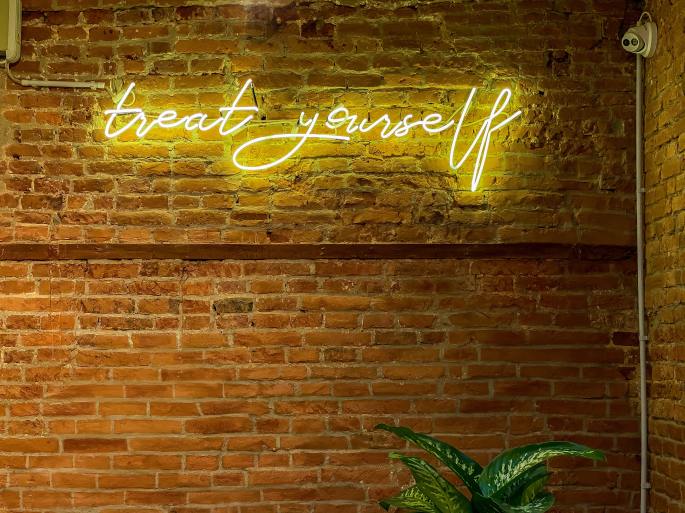 A brick wall with a yellow neon sign that reads "treat yourself." There is a speckled green plant (dieffenbachia) in the bottom of the photo. Photo by Etienne Girardet on Unsplash.