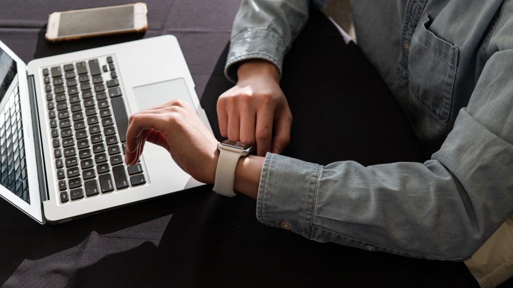 Close up of a person sitting at a table with a laptop and smartphone. The person is looking at their smart watch. 