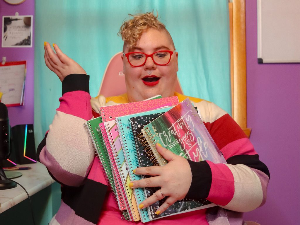 Caitlin is wearing a brightly colored outfit on a pink chair, with a big armful of different notebooks. Their face looks surprised and fun.